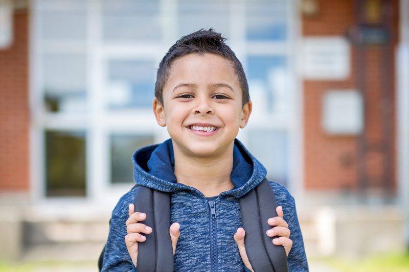 Elementary student smiling at camera and gripping shoulder straps of backpack while standing in front of school building.